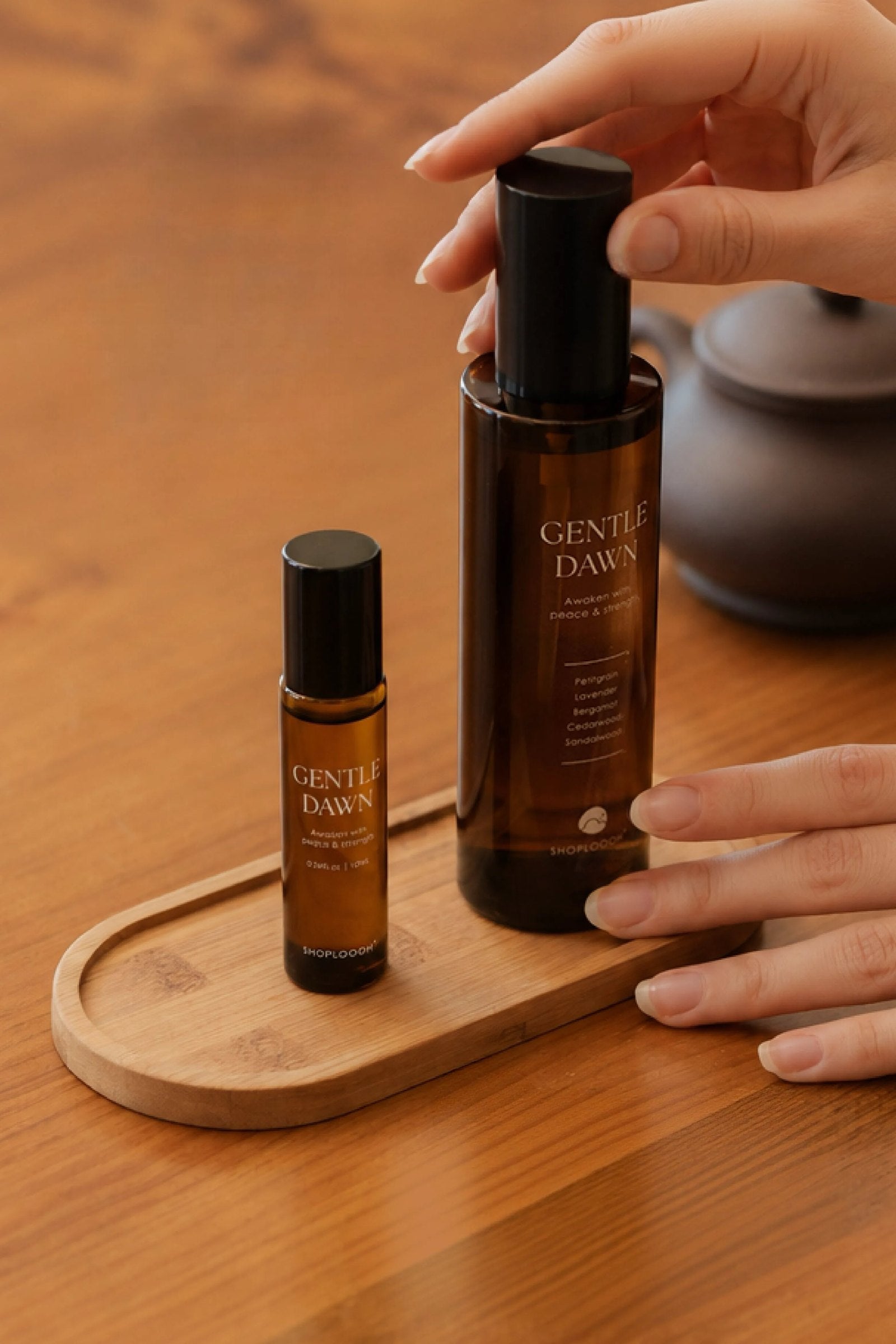 Hands resting on the SHOPLOOOH Gentle Dawn room spray and roll-on, set on a wooden tray beside a clay teapot