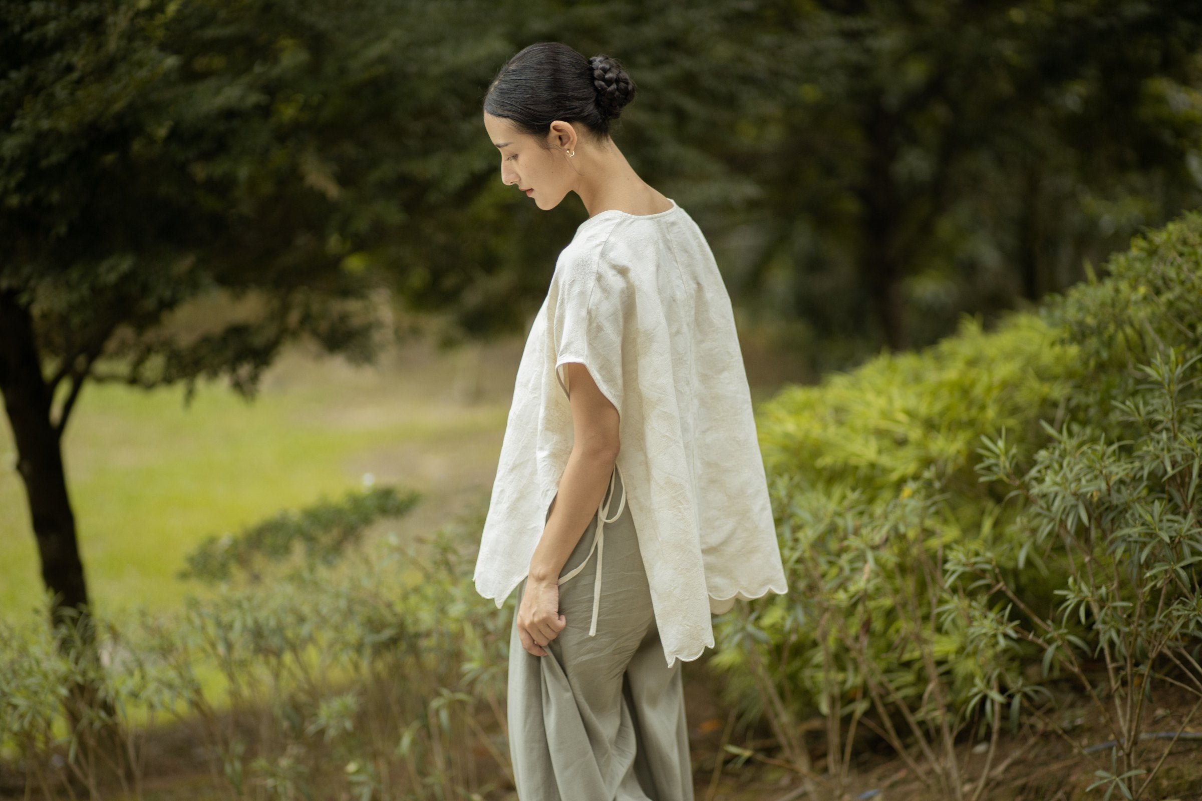 Woman in the SHOPLOOOH Re Collection embroidered scallop top and sage trousers, looking down among the garden greenery